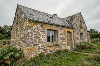 Façade en pierre de granit d'une maison bretonne ancienne à rénover, avec des murs abîmés et une toiture en ardoise dégradée